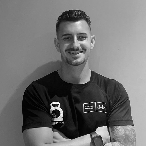 Una persona con cabello corto y barba sonríe con seguridad, con los brazos cruzados. Luce una camiseta con logotipos relacionados con el gimnasio y un reloj inteligente, lo que refleja su firme compromiso con el entrenamiento, sobre un fondo liso.